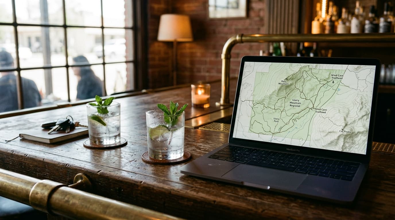 A wooden bar table in a dimly lit Mesa lounge features an open laptop displaying a map of Usery Mountain, next to two half...