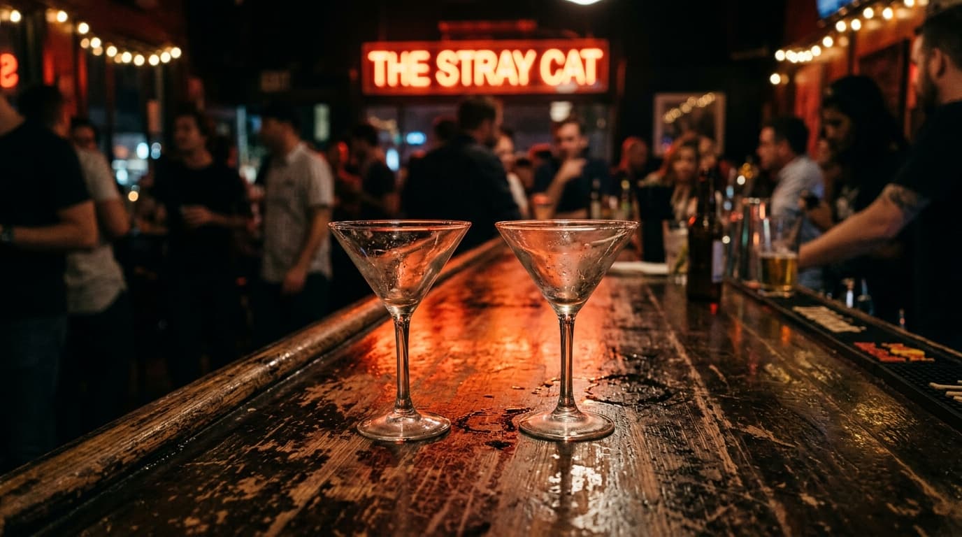 A low-angle shot shows two empty martini glasses resting on a dark, scarred wooden table at The Stray Cat. A red neon sign...
