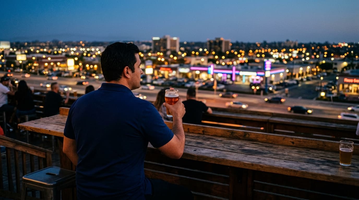 An over-the-shoulder view of a man in a crisp navy polo sitting at a wooden brewery table, glass of craft beer in hand, ov...